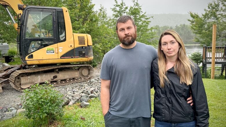 A man with a beard and moustache and a woman with long blonde hair stand in their back yard next to a large piece of heavy equipment.