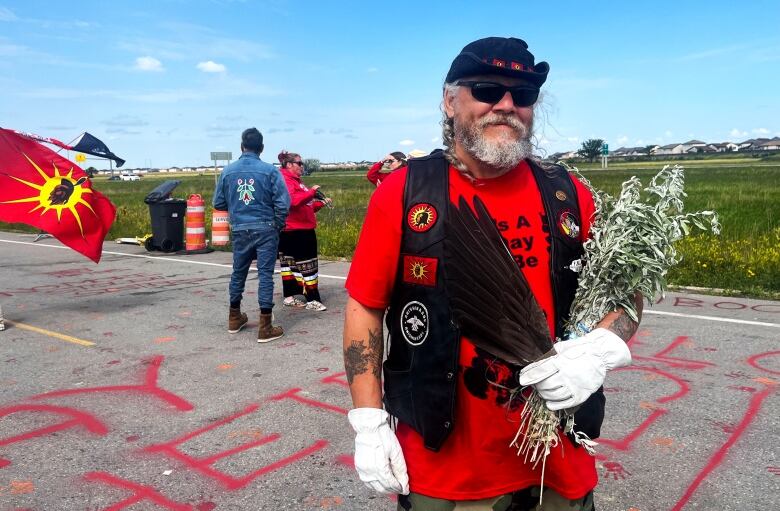 A man in a red shirt and sunglasses holding a feather and safe poses for a photo. 