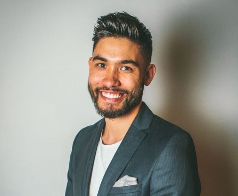 A man with beard and in suit stands in front of a wall smiling.