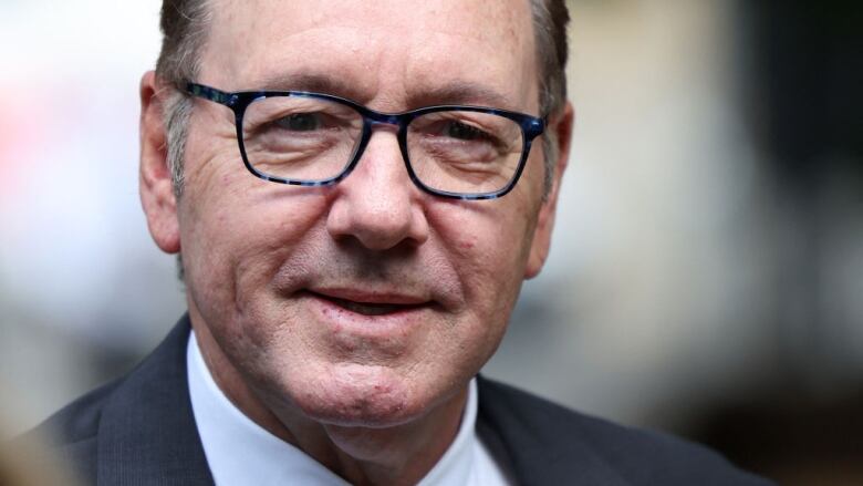 A man wearing blue-frame glasses and a grey suit arrives at a court building in London, England.