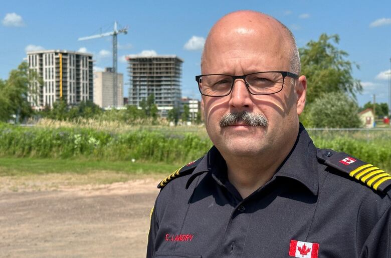 A bald man in a black shirt with his name and a Canada flag in a dirt parking lot with greenery behind him as well as two 15 storey towers under construction. 