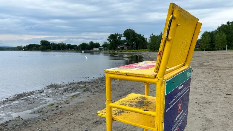 A yellow lifeguard chair with a blue sign on it at a river beach on a cloudy day.