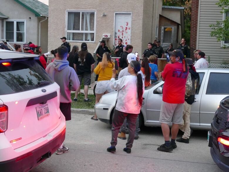 People stand on a street and a sidewalk facing a house with police officers in the front yard and red handprints on the front door.