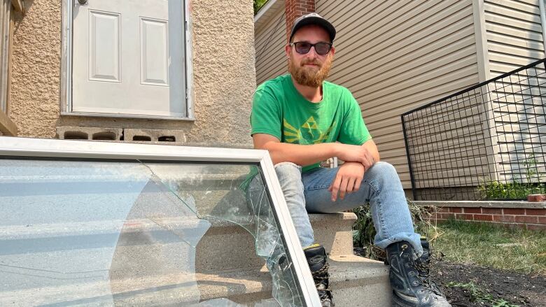 A man in a green T-shirt sits on concrete steps next to a broken window. 