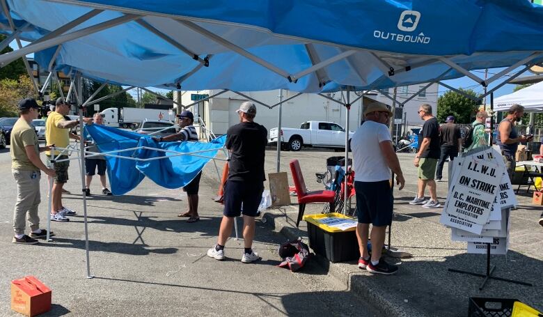 Five men collapse a blue tent canopy in a parking lot as others watch. A stack of picket signs can also be seen.