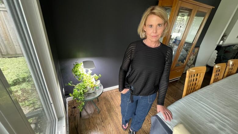 A woman stands in her home, with her hand on a table, staring toward the camera.