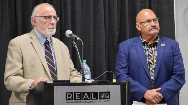 Two men stand near a podium in Regina. Both are wearing suits. 