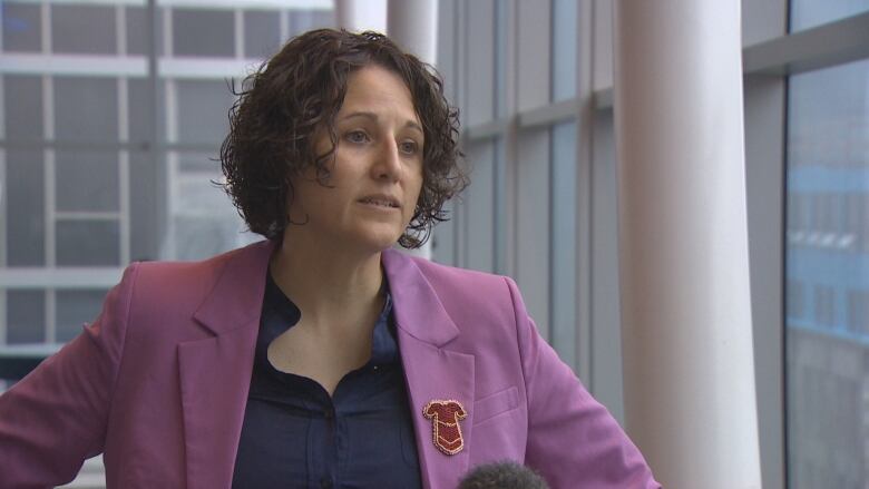 A woman in a purple jacket speaks to the camera.