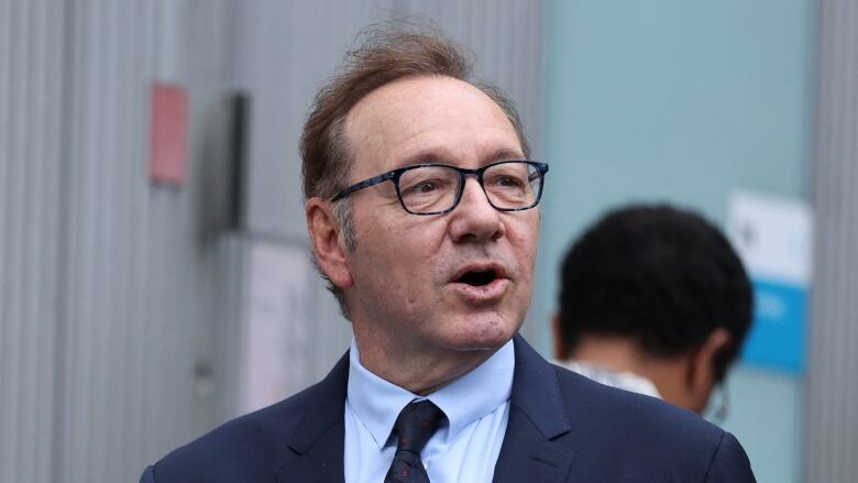 A man in a suit and tie, wearing glasses, appears to be speaking in a photo taken outdoors.