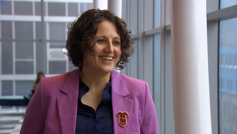 A Mi'kmaw woman is smiling and speaking to a reporter off-camera. She's wearing a purple blazer, with a beaded pin of a red dress on her lapel.