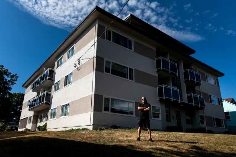 A man stands in front of a three-story apartment building with his arms crossed.