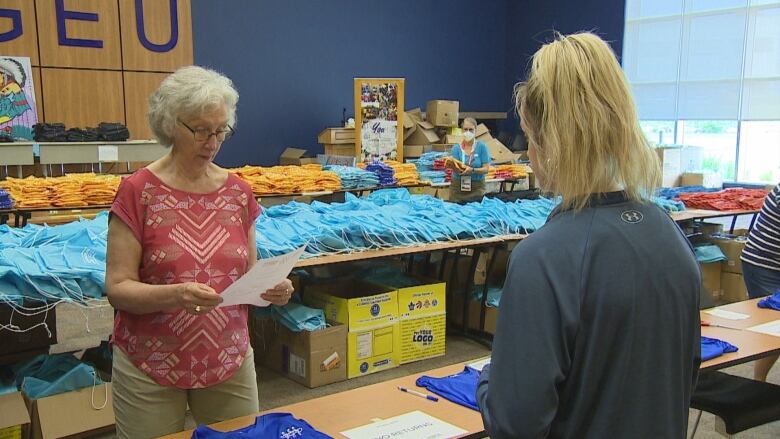 A volunteer in a red patterned T shirt is looking at another volunteer's registration form. Behind her are rows of bags and T-shirts to give out volunteers.