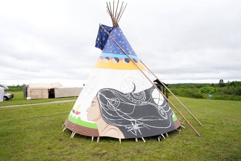 A teepee stands in a field. The cloth features a woman with long flowing black hair which has details of the sun, moon and stars.