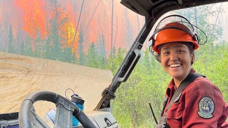 A woman with dark hair smiles for a photo in front of a wildfire. She wears a dusty red button up and a firefighter's helmet.