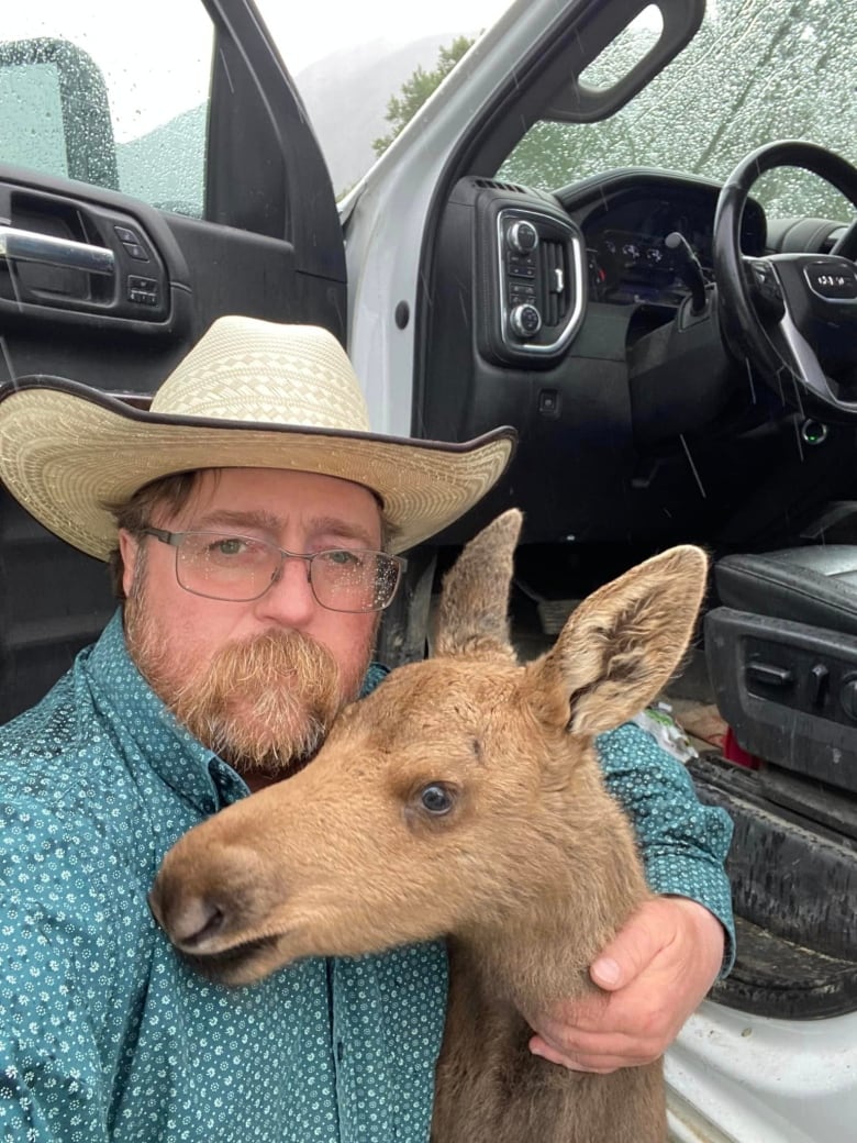 A man with glasses, facial hair, and a wide brimmed hat poses for a selfie with a baby moose.