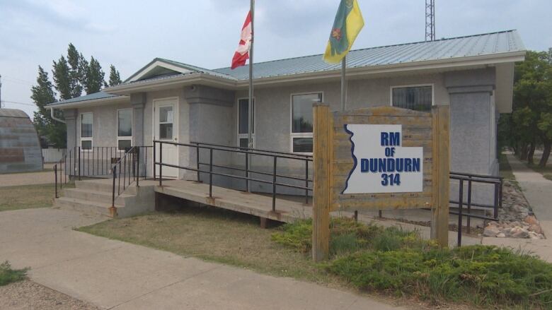 A grey one-storey building with an aluminum roof. Two flagpoles, one flying a Maple Leaf and the other flying a Saskatchewan flag, stand in front of the building. A wooden sign that says, 'RM of Dundurn 314,' also stands in front of the building, over some short shrubs. 