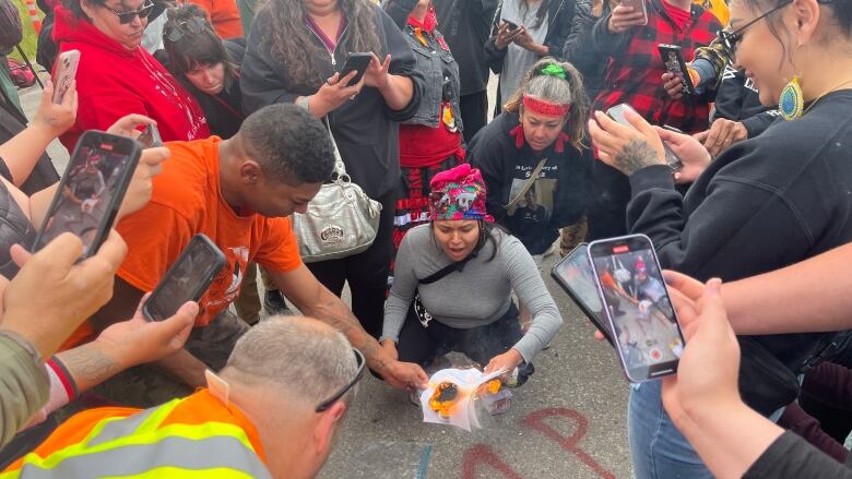 A woman holds burning pieces of paper while people gather around, many recording the act on their phones.