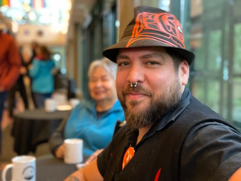 A man in a hat sits in a room with people visible behind him.