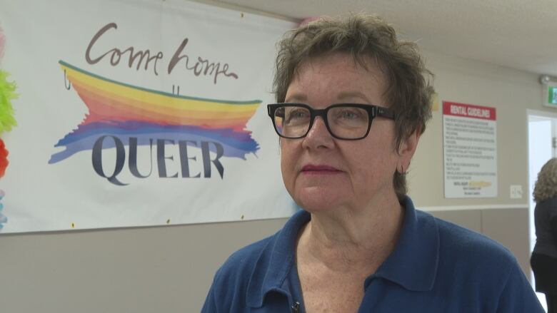 A woman stands in front of a 'Come Home Queer' sign which depicts a rainbow colored fishing boat.