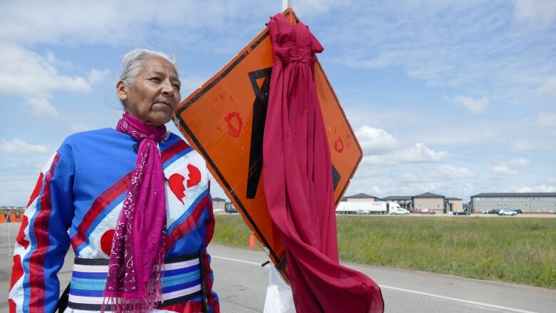 A woman stands on a road next to a sign with a red dress hanging from it.