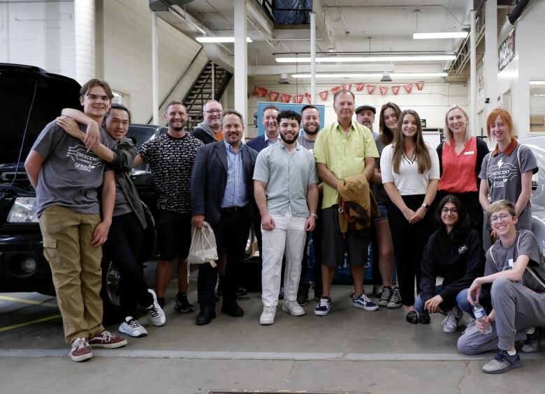 About two-dozen students stand and crouch to pose for a photo in an automotive garage. 