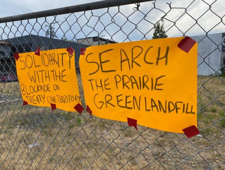 Two signs are taped to a chain link fence. The one on the left says 