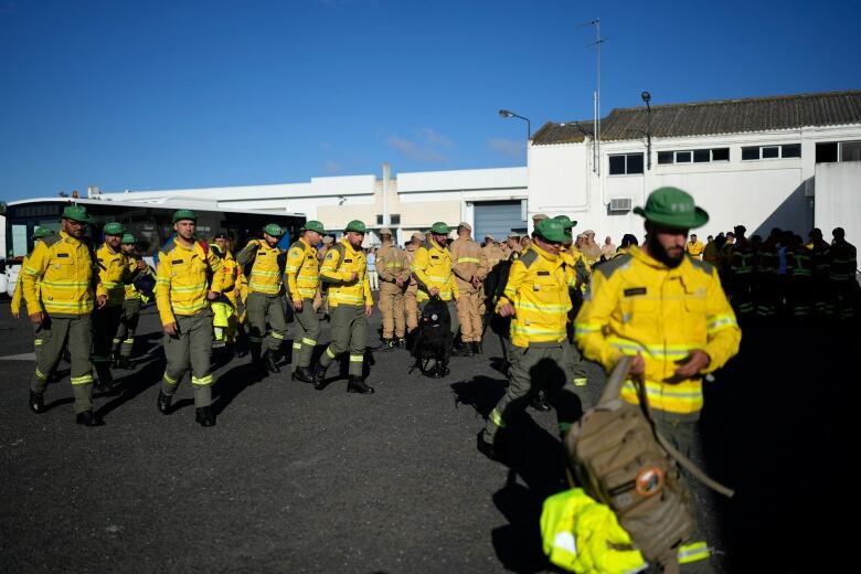 Firefighters in bright yellow uniforms walk across a tarmac at an airport on a sunny day.