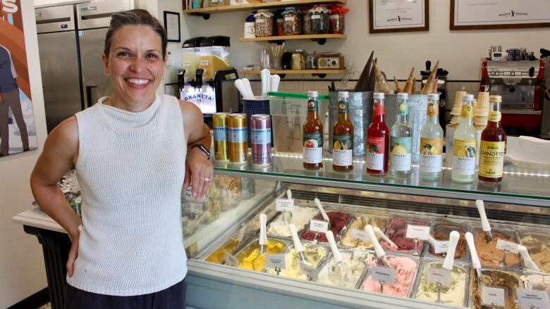 A woman stands in front of a display case of colourful gelato pans. Italian soda bottles and cans line the top of the case.