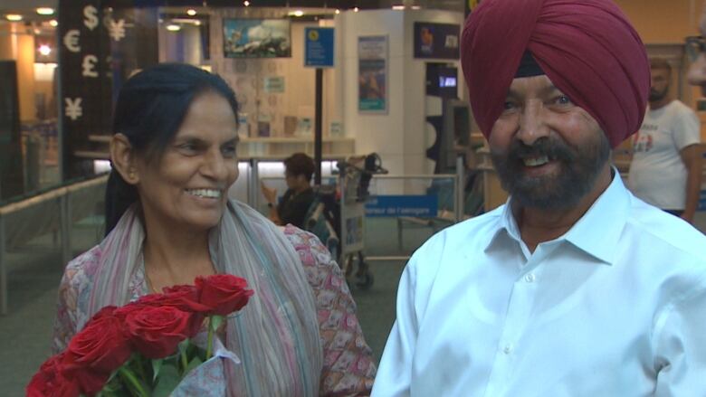 A man and woman laugh at an airport lobby. The woman is holding flowers.