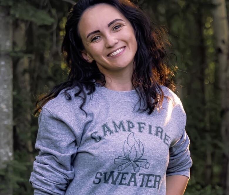 A woman cocking her head and smiling. She wears a grey crewneck that says 