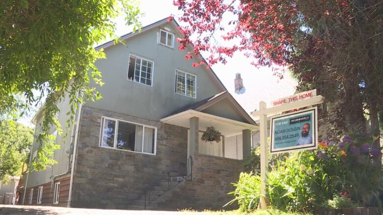 A blue and grey brick home has white pillars and stairs leading up to the door. In front of the house is a sign for Realtor Noam Dolgin with a plaque on top reading, 'Share this home.'