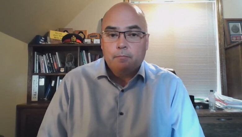 A man wearing glasses and a blue button-up shirt looks forward. Behind him is a bookcase and desk.