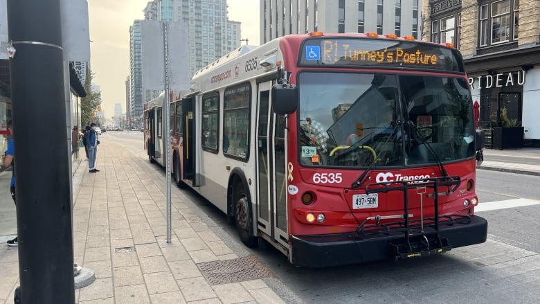 A bus that says R1 Tunney's Pasture stops on a street outside a downtown mall in summer.