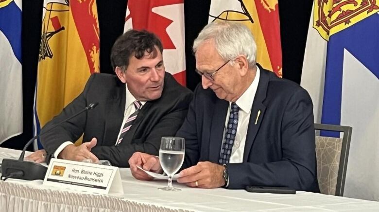 Two men sitting at a table in front of a row of flags from Nova Scotia, New Brunswick and Canada.