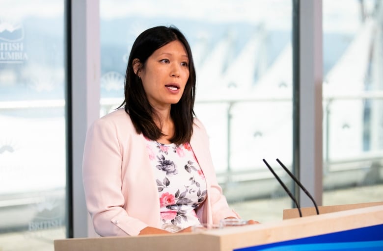 An East Asian woman with shoulder length black hair and pink clothing speaks in front of a lectern.