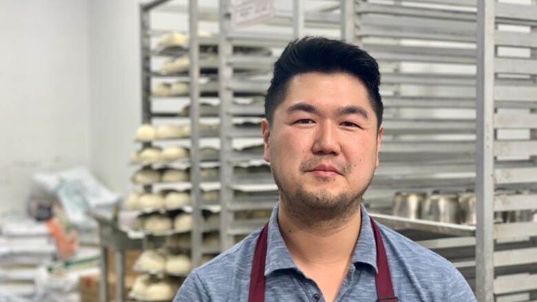 A man wearing an apron stands in front of metal baking shelves loaded with frozen dough in a dim sum shop.