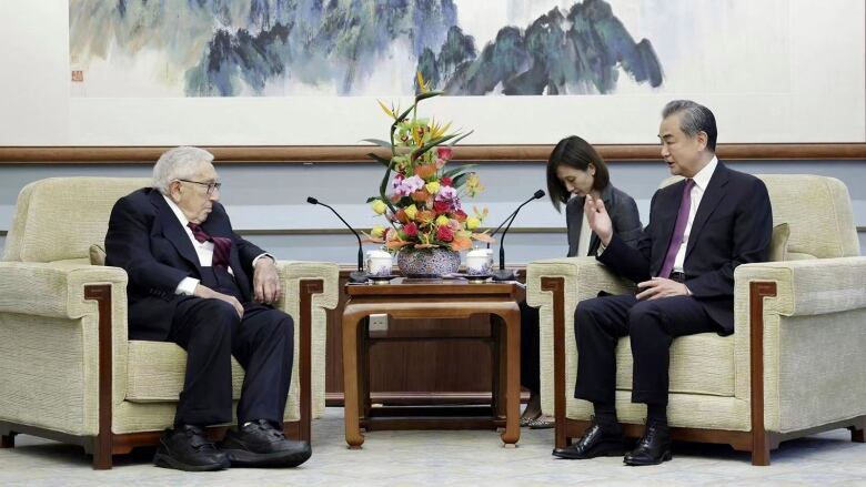 Two men in suits speak to each other while sitting in arm chairs, with a woman shown in the background.