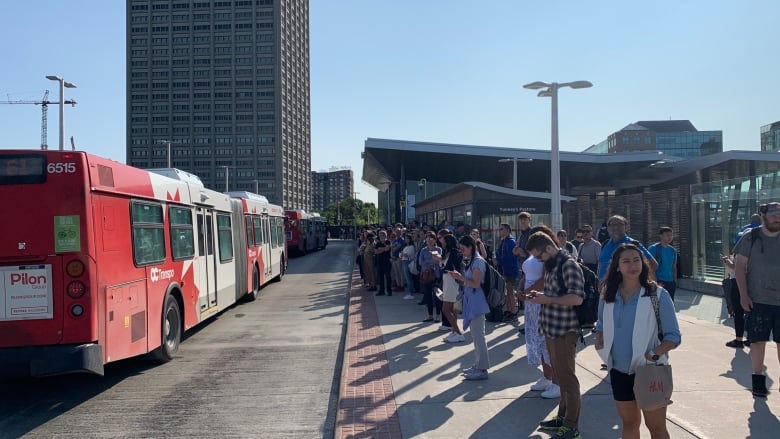 People wait on a crowded bus platform on a summer morning.