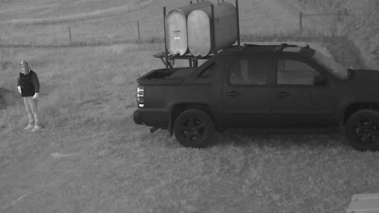 Man wearing a balaclava and jeans outside next to a black Chevrolet Avalanche. 