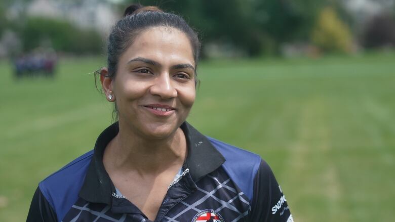 A lady is seen smiling and sporting a dark blue cricket jersey with a high bun.