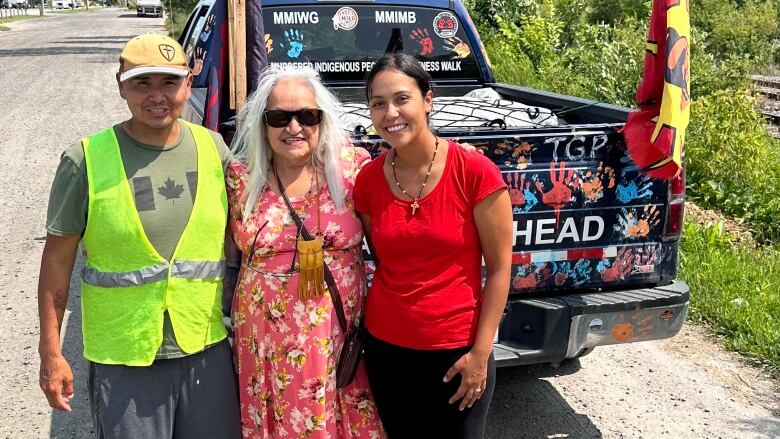 Three people standing at the side of a road with a black pickup truck in the background.