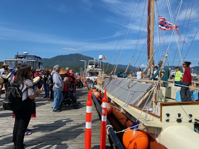 People at a dock near the ocean gather around the crew of a Polynesian canoe on a sunny day. Mountains are visible in the background.