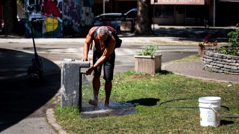 A shirtless man with a backpack wets his feet at a water fountain in a city park. 