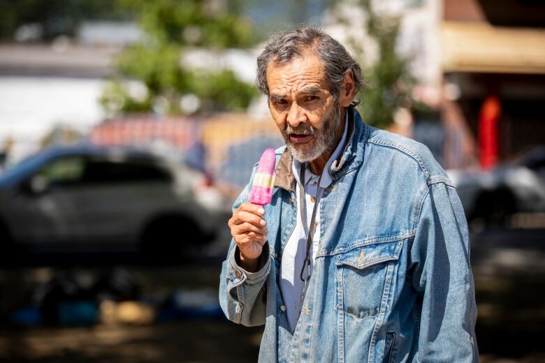 An elderly man with a denim jacket eats a pink popsicle in the sun. 