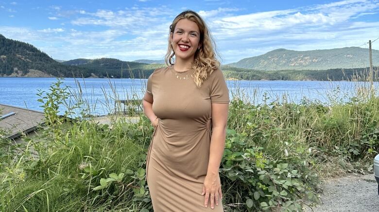 A lady smiles as she poses with a water body and mountains in the background. She's wearing a light brown dress, a dainty necklace and red lipstick.