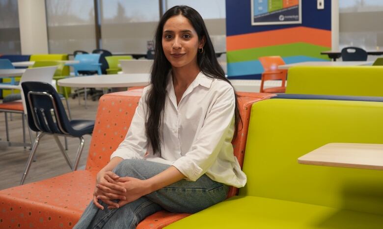 A girl with dark hair is seen in a white shirt and blue jeans with colorful couches and chairs in the background.