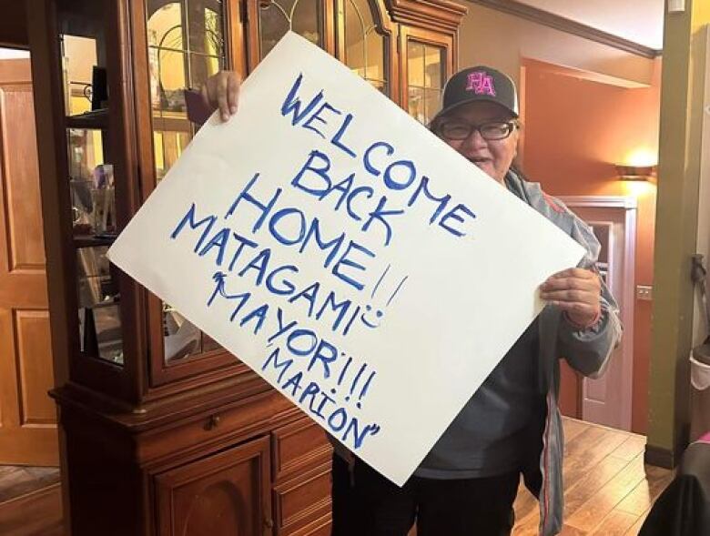 A woman stands holding a hand made sign that says 'Welcome back home Matagami Mayor'. 