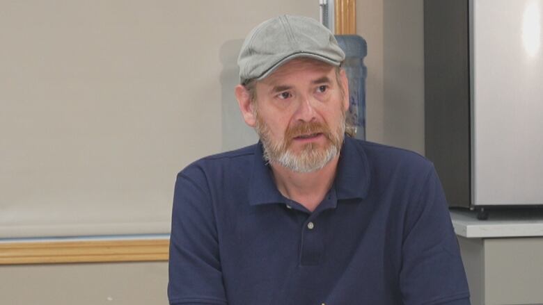 A man with grey beard and a grey hat speaks while sitting behind a desk.