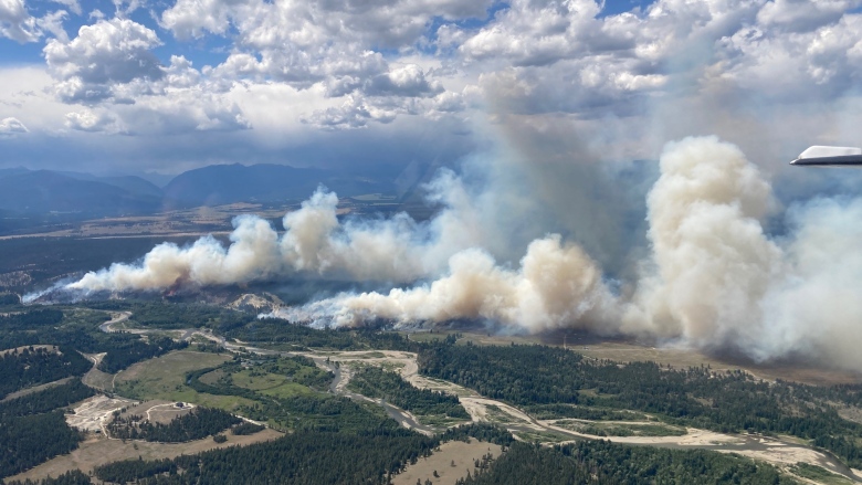 Beige wildfire smoke rises from a fire on a flat, rural area.