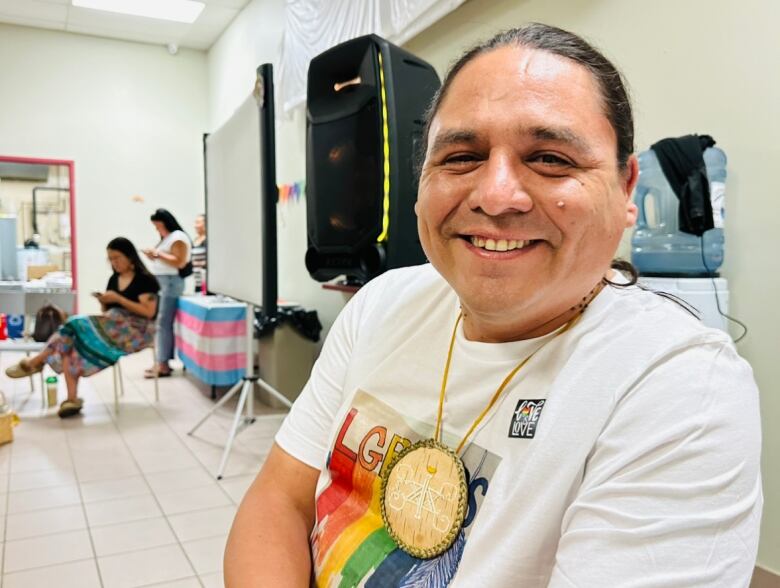 A man with dark hair wearing a t-shirt with a rainbow on it smiles at the camera.